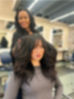 Stylist smiles behind a seated woman with voluminous dark hair in a salon, featuring mirrors and a modern interior.