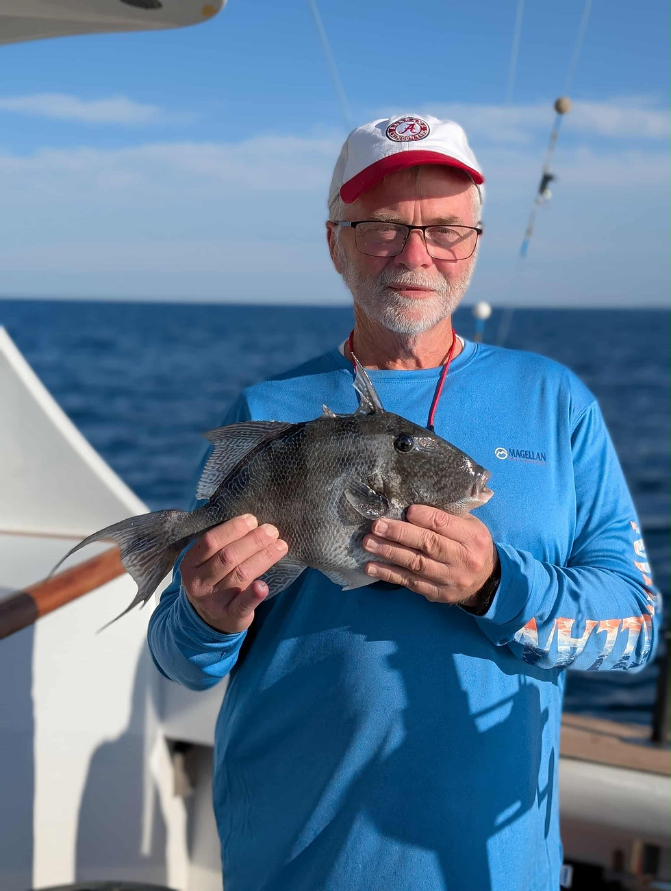Man in blue shirt and red-white cap holds a fish on a boat. Ocean in the background under a blue sky. Looks content.