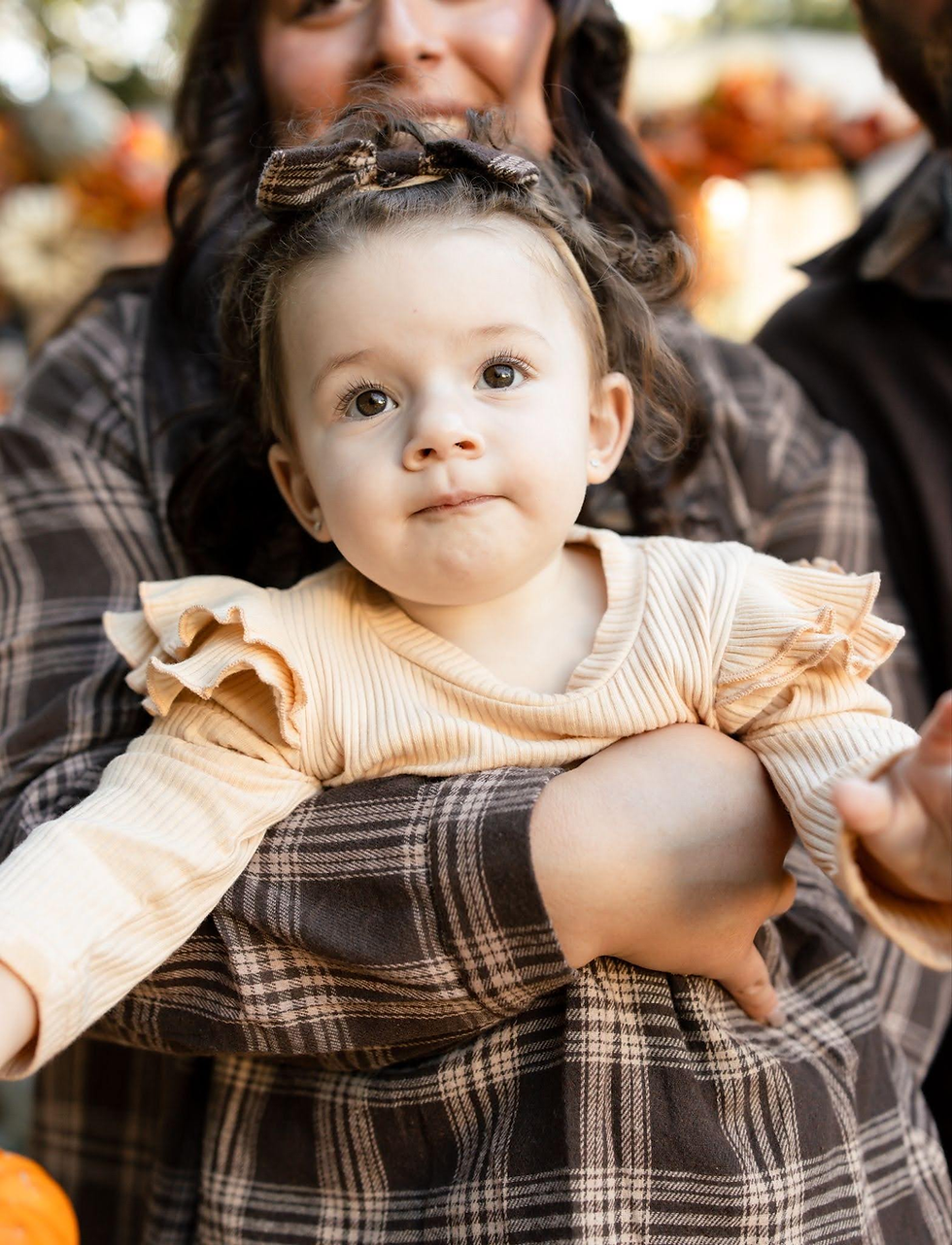 Toddler with plaid bow sits in woman’s arms outdoors, wearing a cream ruffled top, with warm autumn colors and a pumpkin.