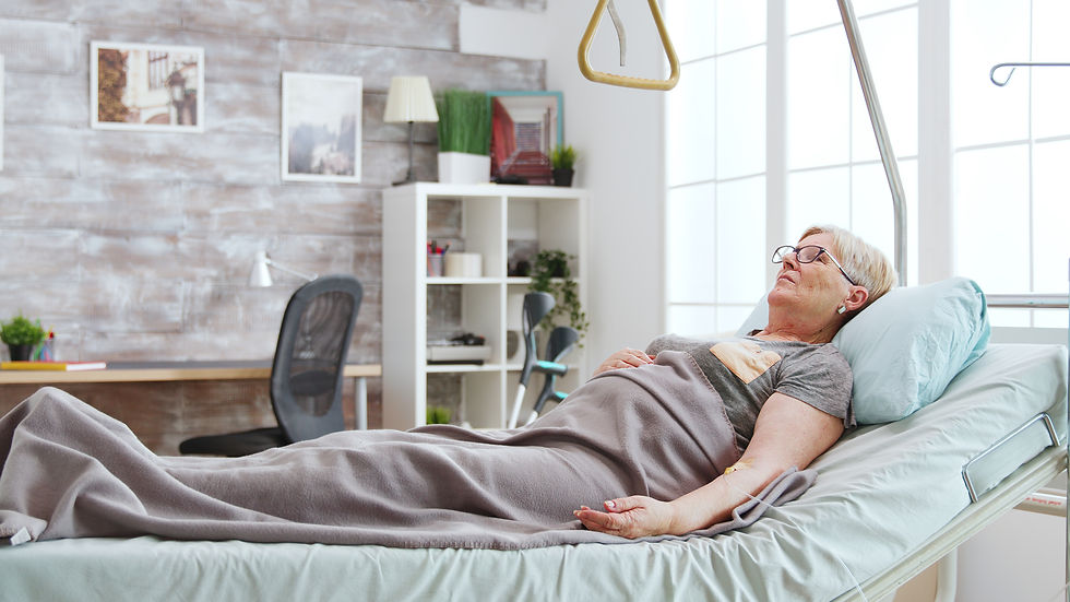 Elderly woman resting in a hospital bed under a gray blanket in a bright room, with a calm expression. Walls display framed pictures.
