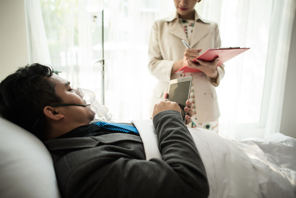 Man in a hospital bed using a smartphone, wearing an oxygen mask. Woman in a white coat with a clipboard stands nearby. Bright room.