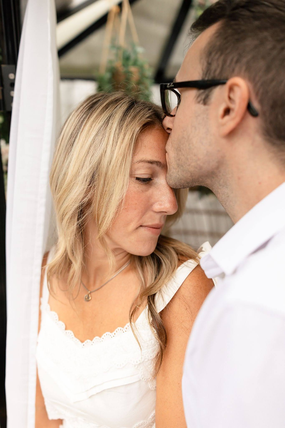 Man kisses a blonde woman’s forehead as she looks down, wearing a white dress; intimate, tender moment indoors with soft decor