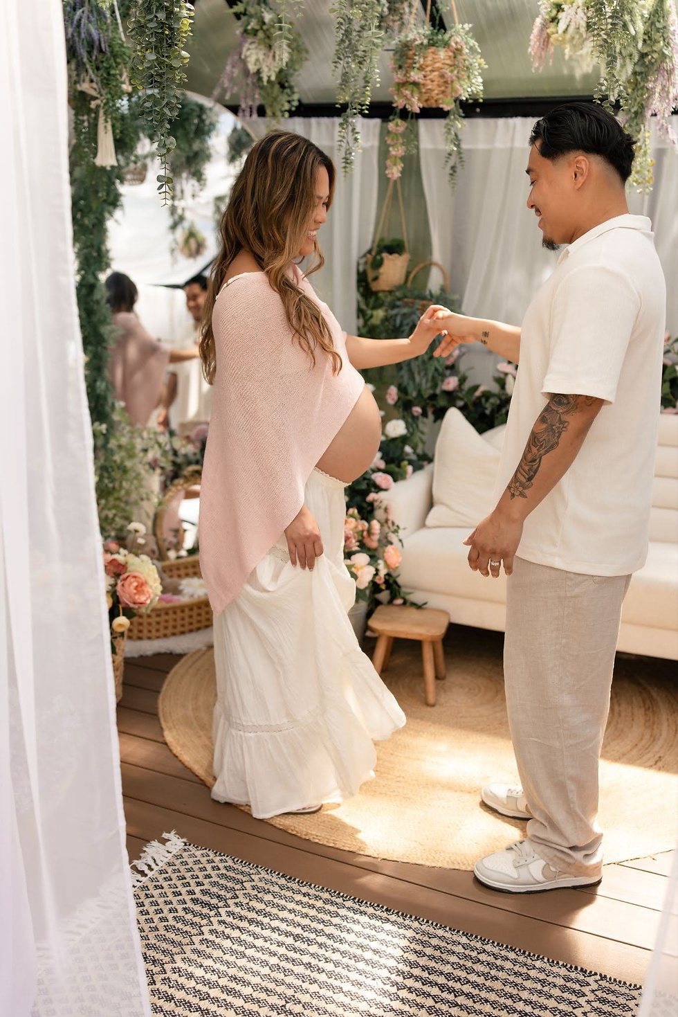 Pregnant woman and man smile and hold hands in a floral-decorated room with hanging greenery and soft white drapes.