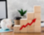 Wooden blocks forming a bar graph with a red arrow pointing up. A piggy bank, coffee cup, and plant are nearby on a desk.