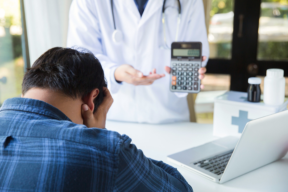 Distressed man covers his face as a doctor holds a calculator in a bright clinic, with laptop and medicine bottles nearby.