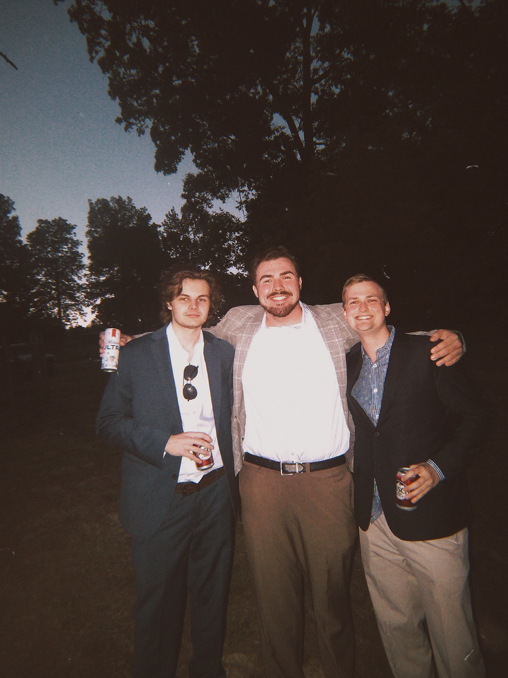 Three friends smiling outdoors at night, holding drinks