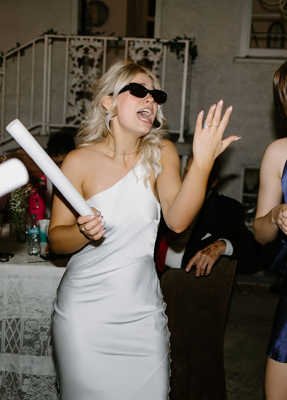 Woman in white dress, sunglasses, holding foam stick at wedding