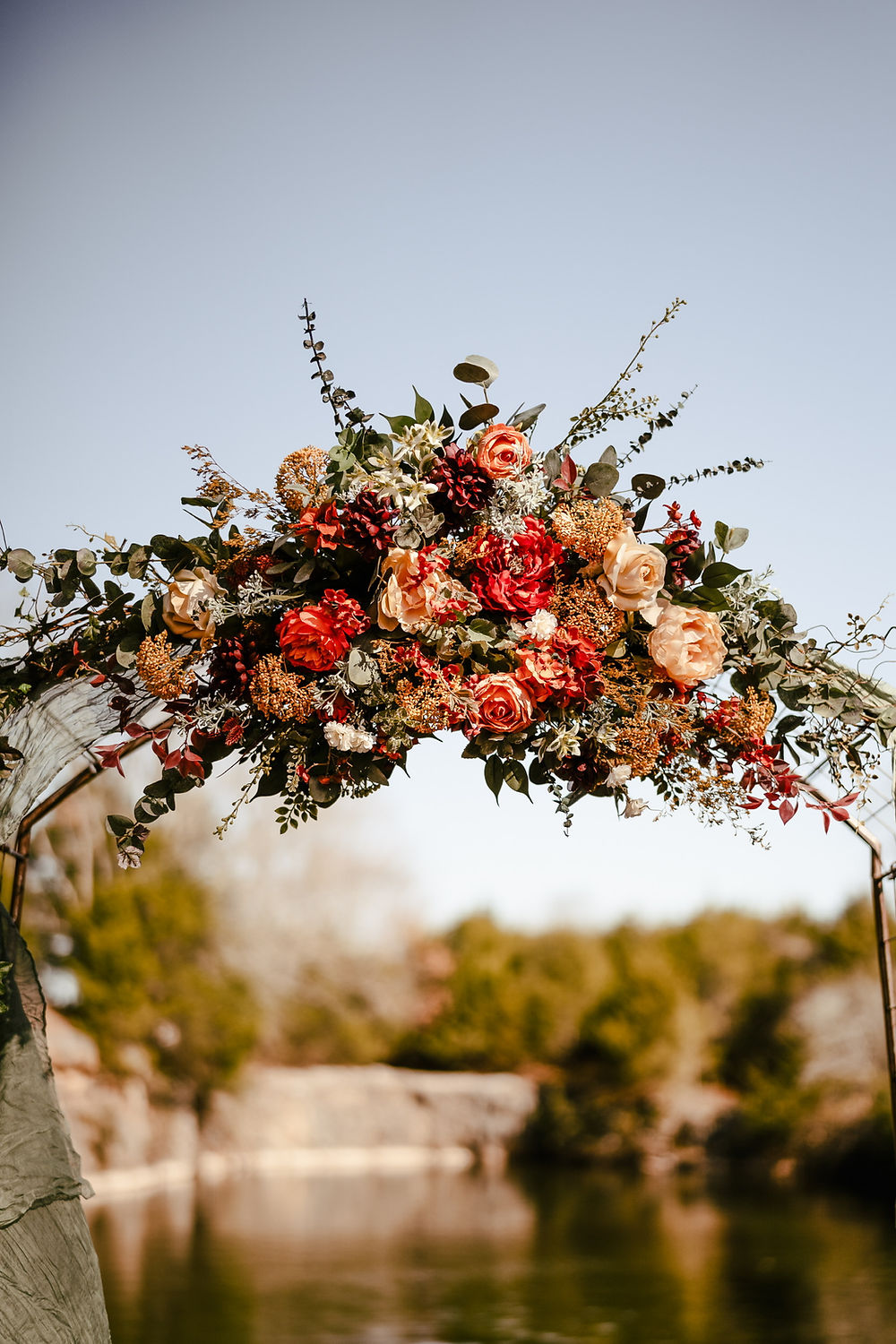 Analea Wedding Co. floral arch with warm flowers over water