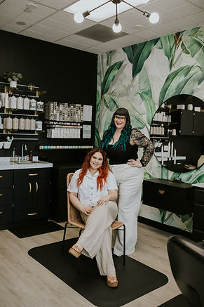 Hairstylist with Copper Hair sitting in a chair with a Hairstylist with Black and Teal Hair Standing