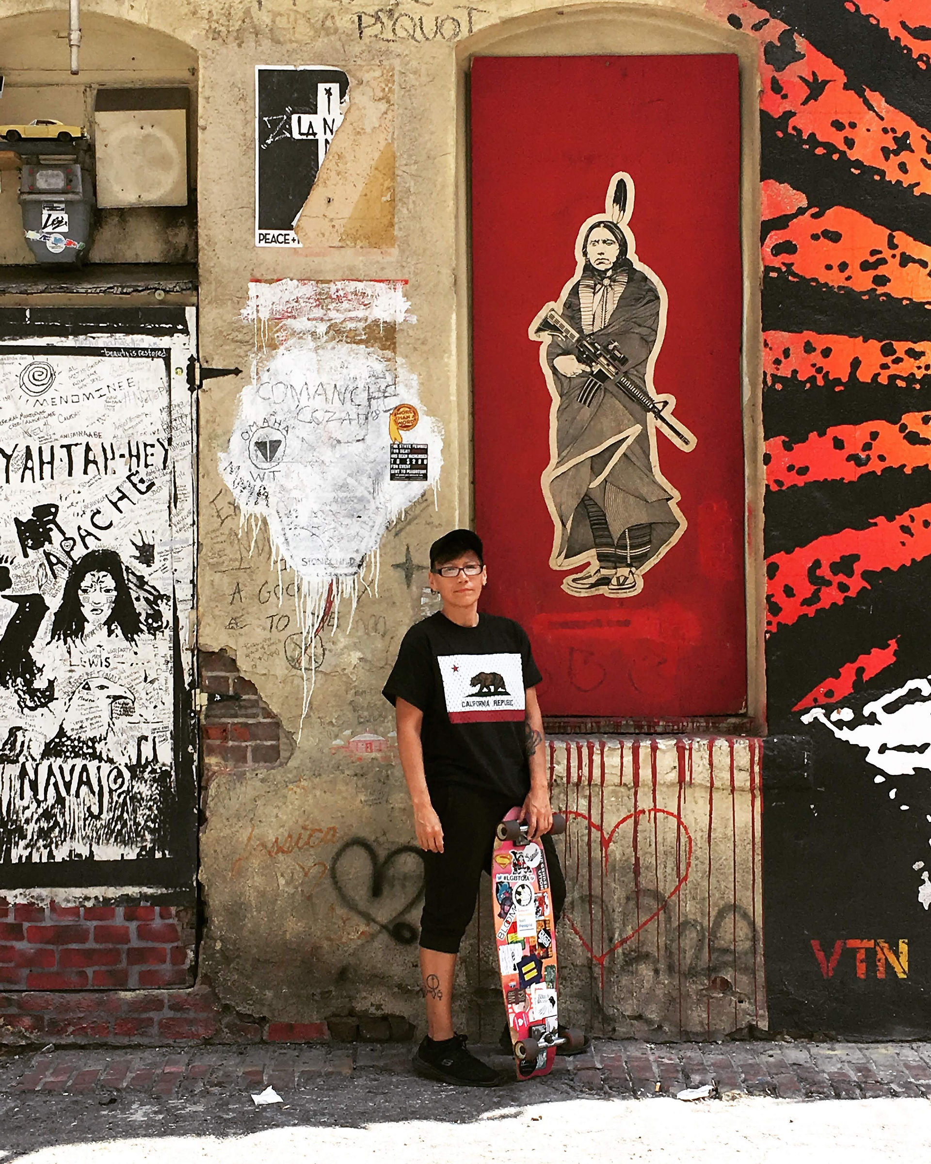 Kit Thomas, Two-Spirit Indigenous artist, standing with a skateboard in Indian Alley in Los Angeles, surrounded by Indigenous murals and street art.