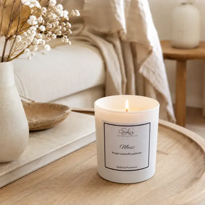 Bougie parfumée naturelle Musc allumée en premier plan dans une pièce à vivre lumineuse, chic et provençale, avec une décoration épurée.