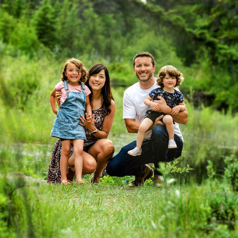 A family of four kneels together on green grass in a lush, wooded outdoor setting. Two adults hold two young children, all smiling toward the camera, surrounded by tall trees and soft natural light. Shot at Griffth Woods Park by Calgary family photographer