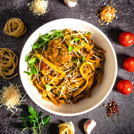 Red pasta with a pork meatball garnished with parmesan cheese, sundried tomato and fresh arugula . Ingredients surround the dish, garlic, noodles, parsley shot by branding food and drink photographer in calgary