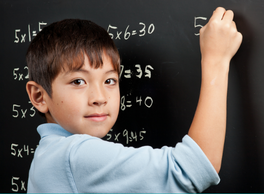 White boy, around age 8, third grader, working on multiplication facts on the black board.