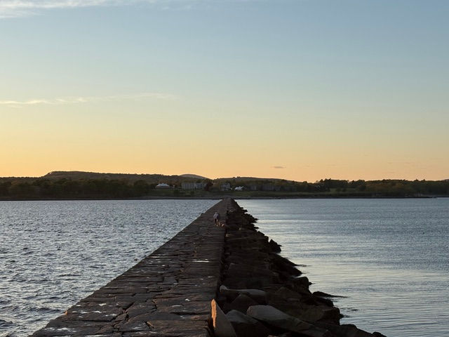 Stone jetty stretches into calm water at sunset, with two tiny figures walking toward the horizon under a pastel sky.