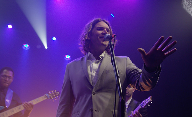 Owner, Sam Ravenna, in a suit at the microphone with ambient stage lighting and a guitarist and bassist behind him.