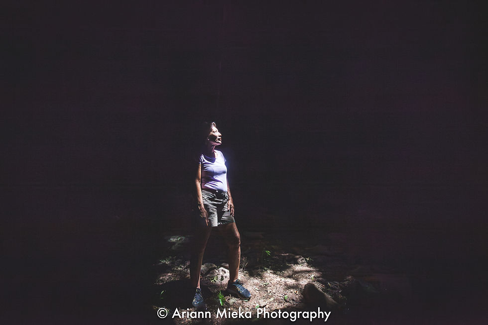 Woman in a spotlight stands on rocky ground in a dark cave, wearing a purple shirt and shorts