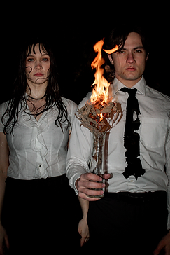 A high-fashion editorial photo from the "Corpurgatory" shoot featuring sharp lighting and professional styling. A woman and man in office wear. The woman is drenched and the man has a burnt tie and is holding a plant that is burning.
