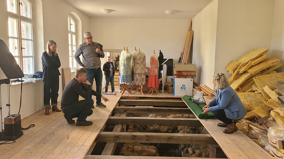 Experten stehen und knien in einem Museumsraum mit geöffnetem Holzboden und betrachten einen freigelegten historischen Ofenbefund im Boden. Im Hintergrund stehen Schaufensterpuppen mit Kleidern sowie Baumaterialien, was auf laufende Planungen und Untersuchungen im Schloss Meyenburg hinweist.
