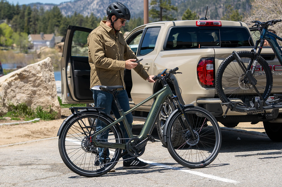 Man in helmet using phone near a green bike and a beige truck. Open door and bike rack visible. Mountainous, sunny background. Calm mood.