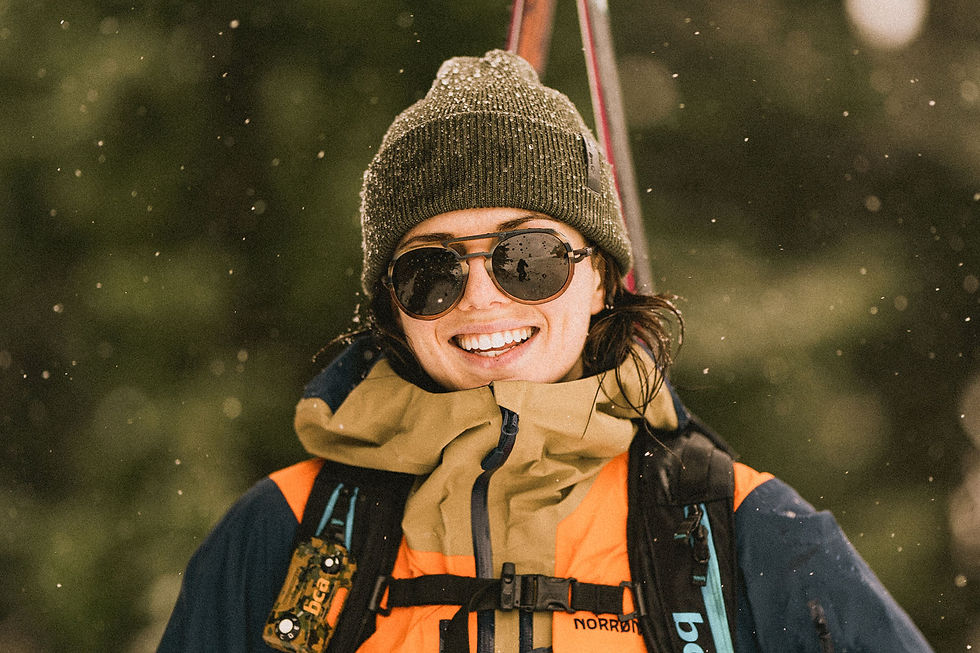 Smiling person in winter gear, sunglasses, and a beanie, surrounded by falling snow outdoors. Orange and green jacket with "Norr" visible.