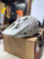 White helmet on a cardboard box with the logo "Sweet Protection." Blurred background shows colorful snowboard designs.