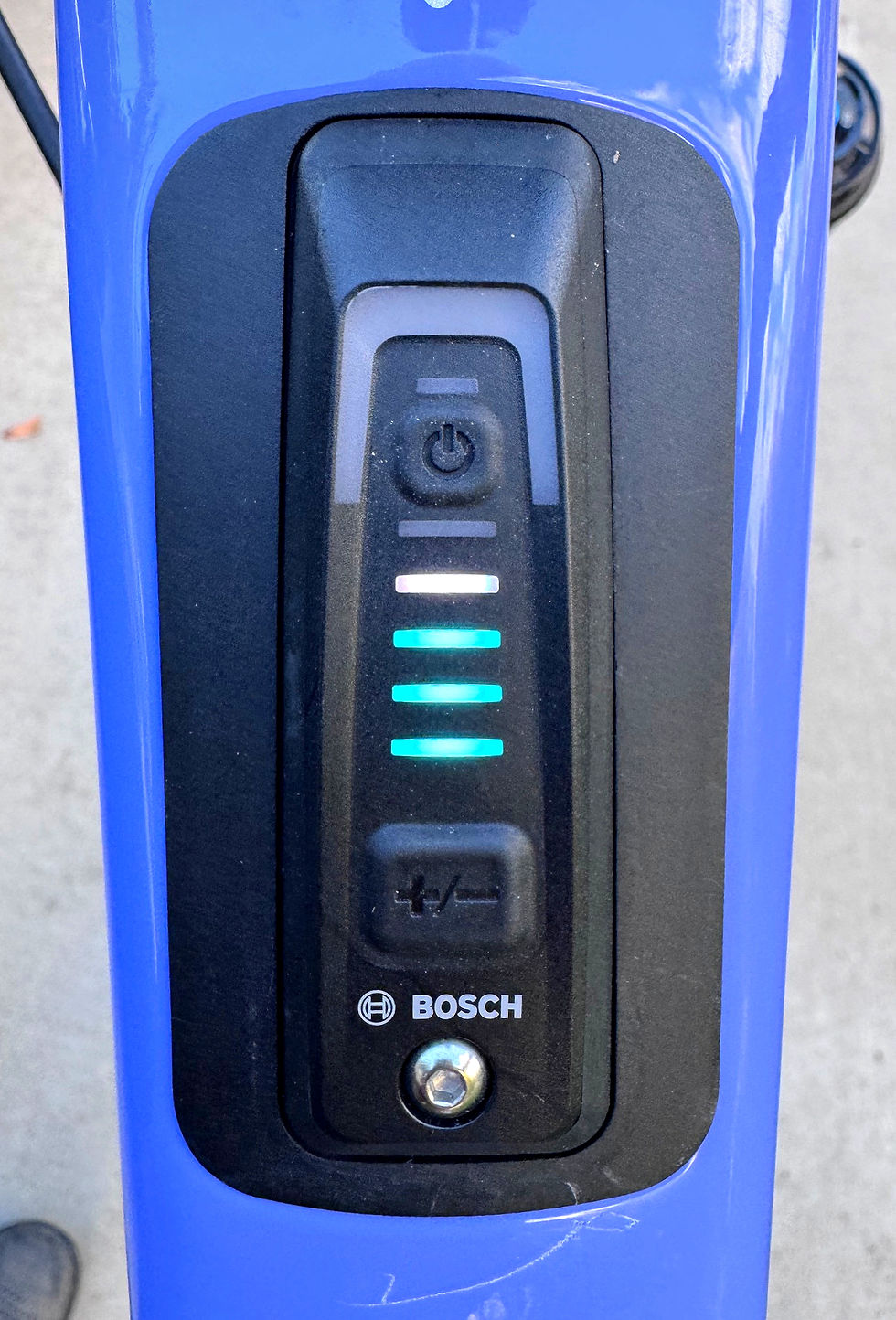 Close-up of a Bosch battery indicator on a blue e-bike frame. Four green lights displayed, suggesting charge level. Concrete background.