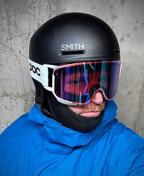 Person in a blue jacket wearing a black helmet and reflective ski goggles against a concrete wall. Helmet reads "SMITH," goggles "POC."