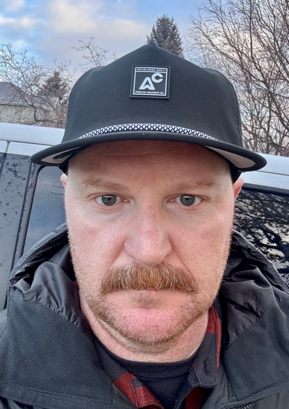 Man in a black cap with logo, unsmiling outdoors. Grey sky and bare trees in the background. Wearing a dark jacket.