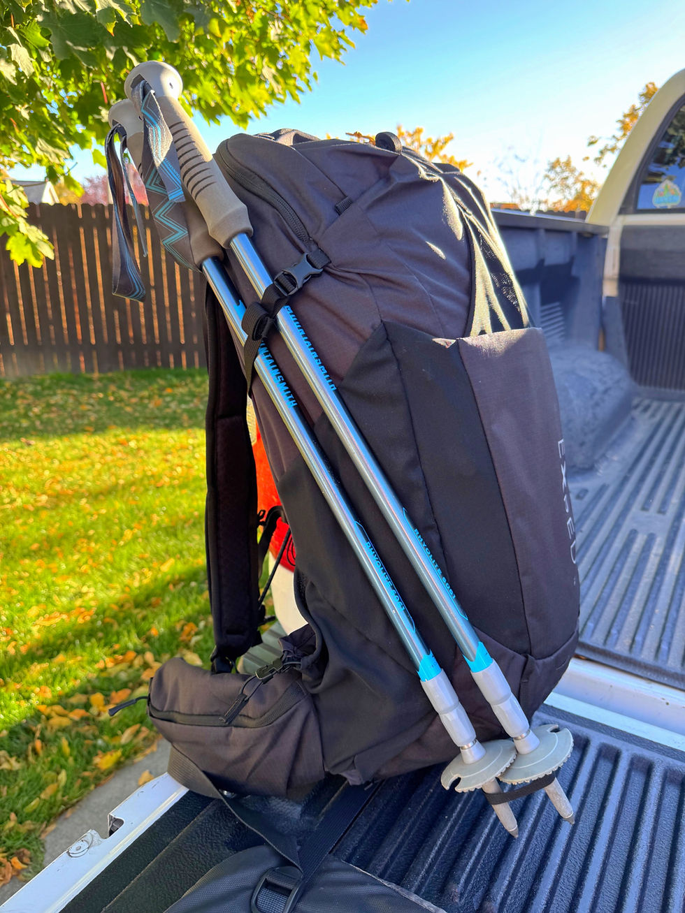 Black backpack with blue trekking poles attached, resting on a truck bed. Sunlit green grass and wooden fence visible in the background.