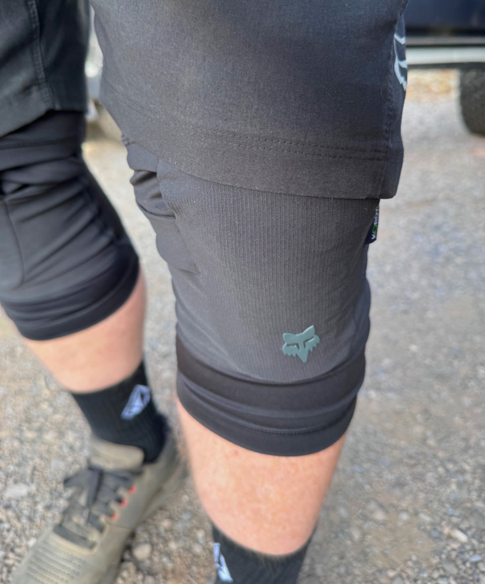 Close-up of a person wearing black shorts with a fox logo, black socks, and gray sneakers standing on gravel.