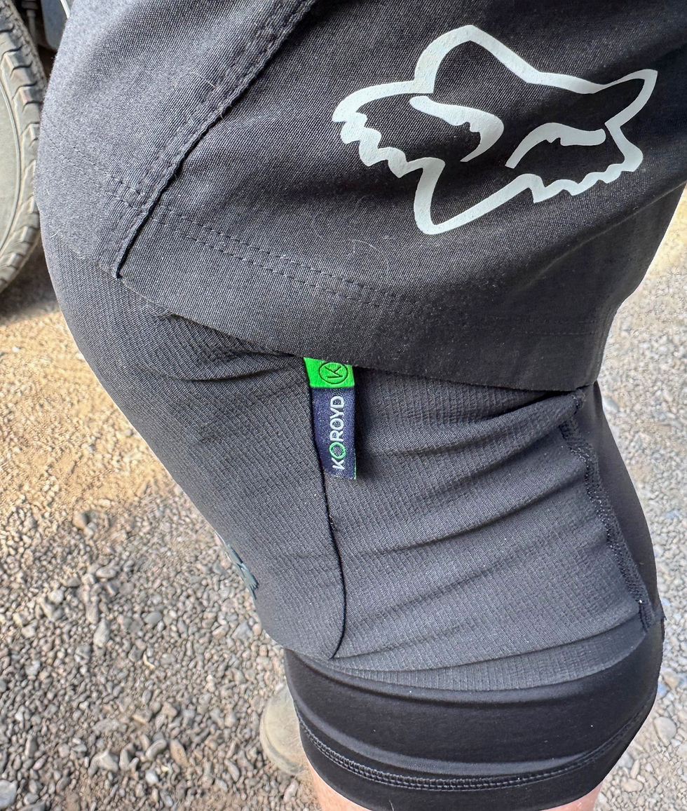 Close-up of a person wearing black shorts with a fox logo and a green KOROYD tag. Background shows a rocky ground and a tire.