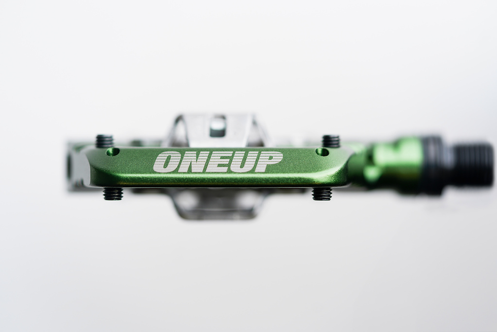 Green metal bicycle pedal with "ONEUP" text, screws visible, against a blurred white background.