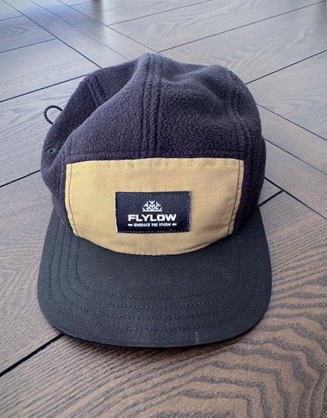 Black and tan cap on wood floor. "FLYLOW" text on front patch. Cap is made of soft material with a black brim.