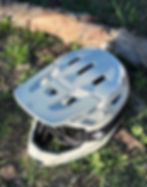 A white bike helmet with goggles rests on grass next to a log. "Sweet Protection" logo visible. Sunlight casts shadows.