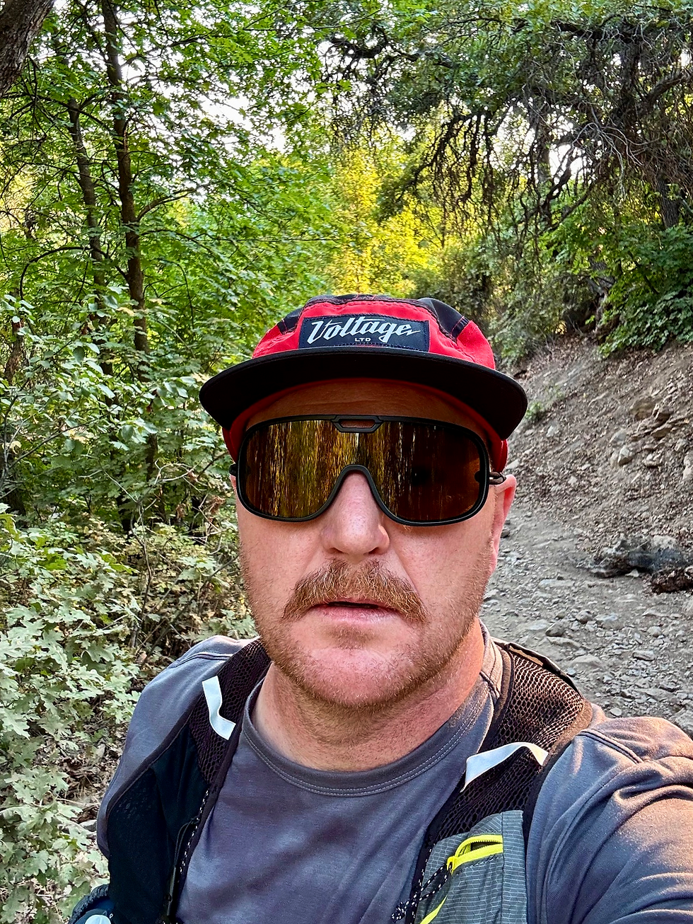 Man with a mustache wearing sunglasses and red cap labeled "Voltage," standing on a forest trail surrounded by green foliage.