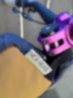 Bicycle handlebar with a purple mount, tan bike bag labeled Oveja Negra Adventure Cycling Gear, against a wooden fence background.