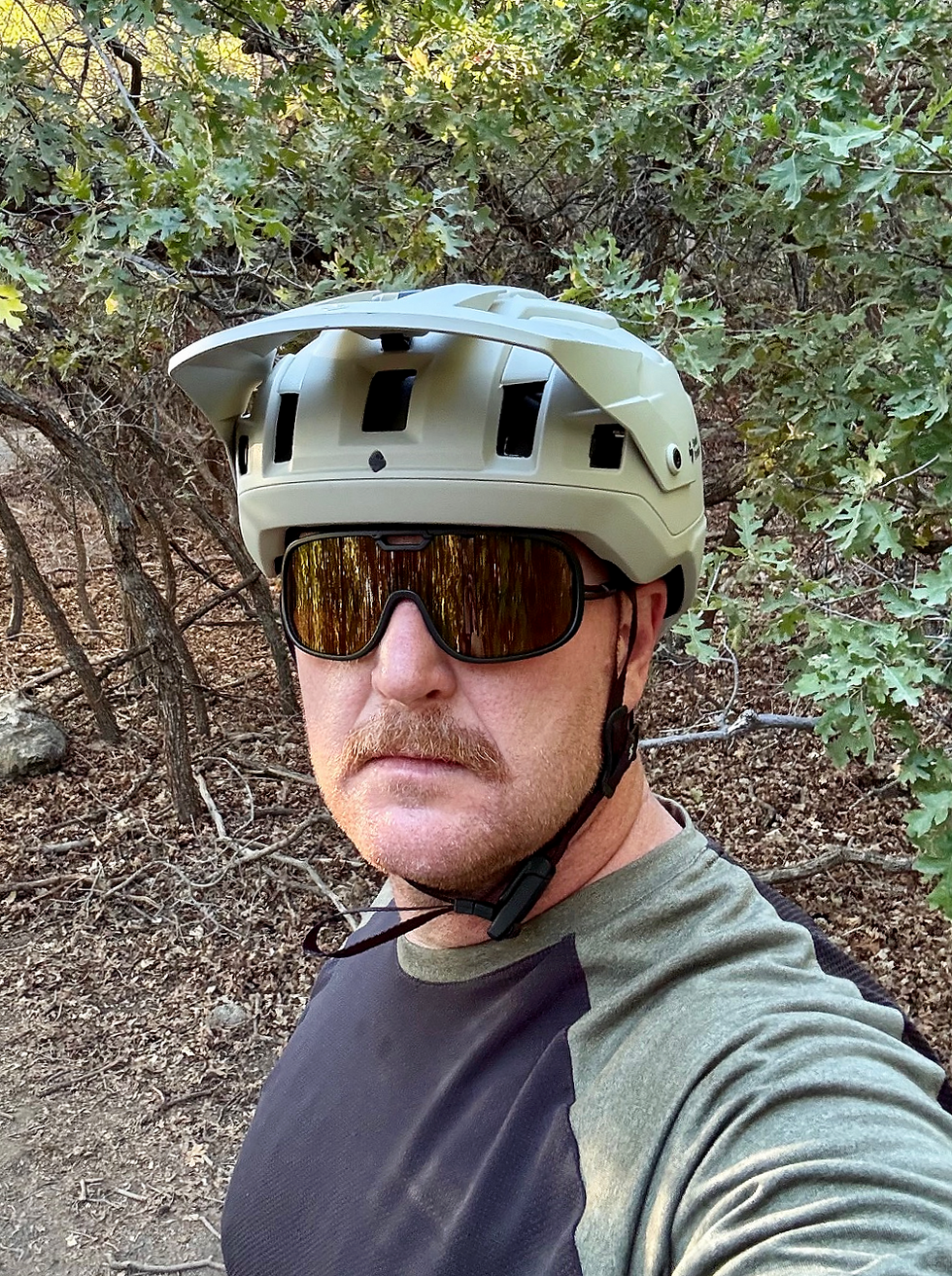 Man in a trail setting, wearing a bike helmet and sunglasses, with a serious expression. Background has green foliage and brown leaves.
