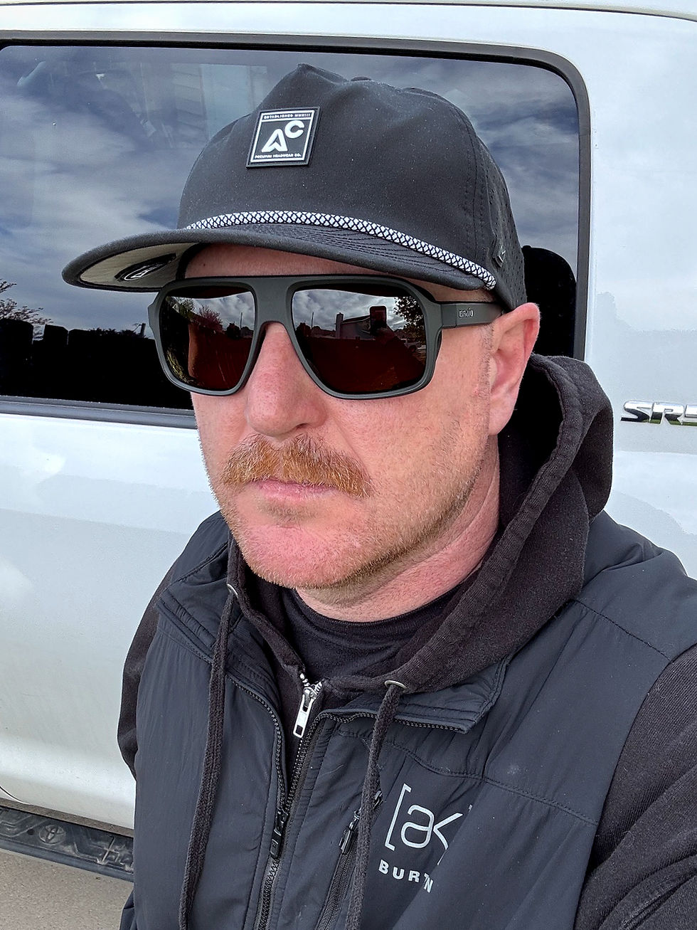 Man wearing sunglasses, black cap with "AC" logo, and black jacket stands in front of a white vehicle. Reflective windows show a cloudy sky.