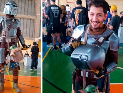 Cosplayer de Mandaloriano sorrindo em convenção, com Grogu no cinto, em ginásio movimentado.