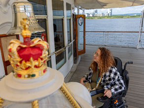 Woman in a wheelchair on a yacht deck, smiling near a crown-topped bell; river and city skyline beyond.