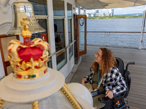 Woman in a wheelchair on a yacht deck, smiling near a crown-topped bell; river and city skyline beyond.