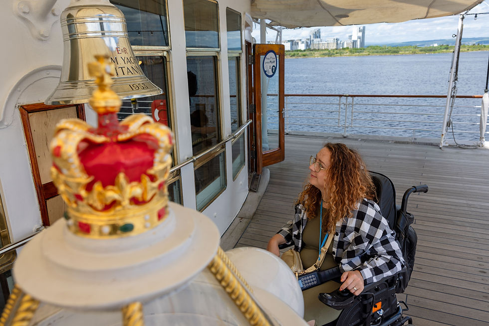 Woman in a wheelchair on a yacht deck, smiling near a crown-topped bell; river and city skyline beyond, with M.Yacht Titan 1953 on bell