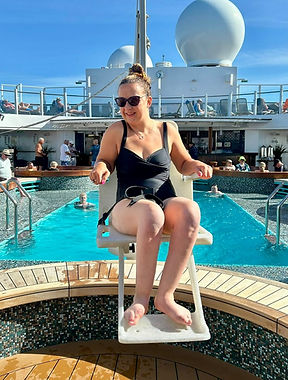 Carrie-Ann Lightley sits on a pool hoist on a cruise ship deck, wearing a black swimsuit and sunglasses. She is positioned above the water at the edge of the pool, with other guests swimming and relaxing around her under a bright blue sky.
