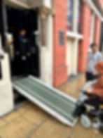 Person in wheelchair using a ramp to enter a building with red walls; a man observes and a staff member holds the door open. Signs visible.