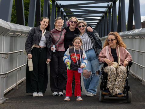 Group photo of six people on a ramped pedestrian bridge, including one wheelchair user and one child standing at the front. Everyone is smiling at the camera, with metal bridge railings and grey beams framing the walkway behind them.