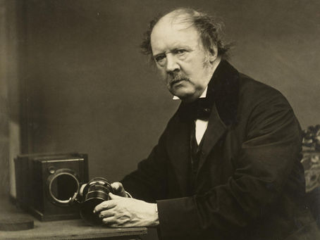A seated portrait of William Henry Fox Talbot, dressed in formal 19th‑century attire, holding a large camera lens in both hands. An early wooden box camera rests on the table beside him. - William Henry Fox Talbot, the British inventor of the negative-positive photographic process, photographed around 1864 by John Moffat. This portrait is held in the National Galleries of Scotland collection. Image © National Galleries of Scotland (used for educational purposes).