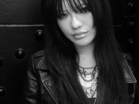 Black-and-white portrait of a young woman with long dark hair and sunglasses resting on her head. She is wearing a black leather jacket and layered necklaces while leaning against a dark metal wall with visible rivets. Her expression is calm and reflective, creating a moody, contemporary street photography aesthetic.