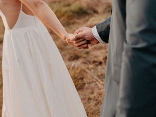 Mariée tenant la main de son partenaire dans un paysage naturel, portant un peigne floral en fleurs en papier rose et corail dans ses cheveux, accessoire de coiffure créé par une fleuriste mariage à Nantes.