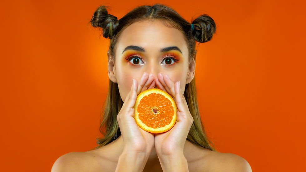 Woman with bold orange eyeshadow holding half an orange in front of her mouth against a bright orange background